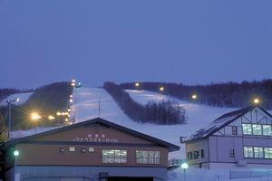薄暗い中、真っに白に雪が積もっているスキー場に電気が灯り、手前には建物が2棟立っているウナベツスキー場の写真