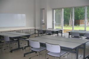 向い合わせの机に椅子が設置されており、左側の壁にホワイトボードがある実習室1の写真
